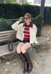 Hvit denimjakke med brun krage, rødstripet polo skjorte, denimshorts, brune støvler til knærne, sittende på en benk i tre, plantebakgrunn.