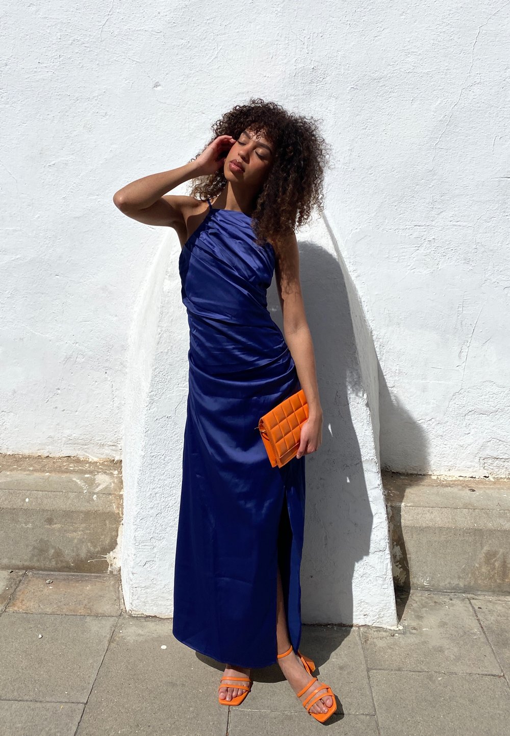 Navyblauwe satijnen jurk met gedrapeerd ontwerp en hoge split tot boven de dij. Oranje gewatteerde clutch en bijpassende sandaaltjes met bandjes. Achtergrond: effen witte muur.