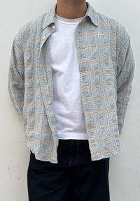 Cămașă texturată de culoare albastră deschisă cu un model în carouri, cu deschidere frontală și închidere cu nasturi, purtată peste un tricou alb și blugi negri.