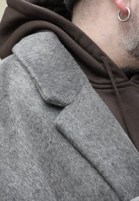 Homme portant un manteau en laine gris par-dessus un sweat à capuche marron foncé, avec une boucle d'oreille argentée visible à l'oreille.