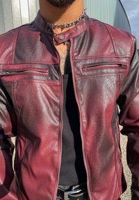 Homme portant une veste en cuir bordeaux déboutonnée par-dessus une chemise noire, avec un collier en chaîne cloutée argenté et un tatouage visible sur la poitrine.