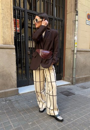 Mujer con blazer marrón oversize, pantalones de pierna ancha a cuadros, grandes gafas de sol, bolso de cintura burdeos, de pie en una acera urbana.