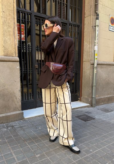 Mujer con blazer marrón oversize, pantalones de pierna ancha a cuadros, grandes gafas de sol, bolso de cintura burdeos, de pie en una acera urbana.