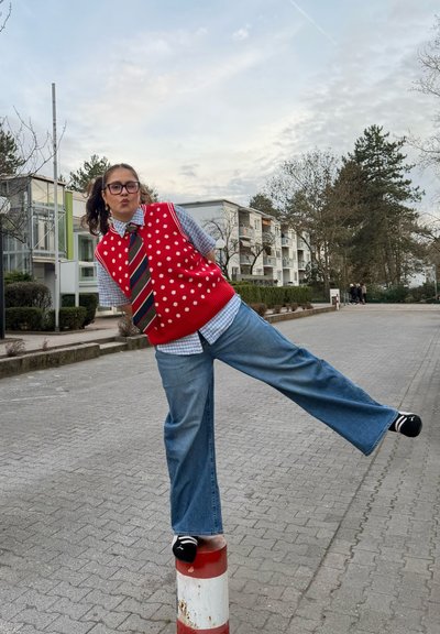 Joven con chaleco rojo de lunares, corbata a rayas y gafas, que se equilibra sobre una pierna encima de un bolardo rojo y blanco en una calle pavimentada.