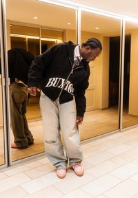 Άνδρας με άνετα φαρδιά ρούχα και ροζ παπούτσια κοιτάζει προς τα κάτω σε ένα δωμάτιο με ντουλάπες με καθρέφτες και πλακάκια στο πάτωμα.