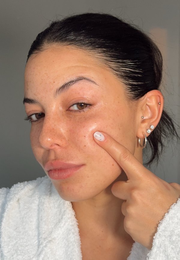 Femme à la peau mate avec des taches de rousseur, portant un peignoir blanc, pointant vers sa joue. La peau semble fraîche et légèrement brillante. L'ongle présente un dessin simple.