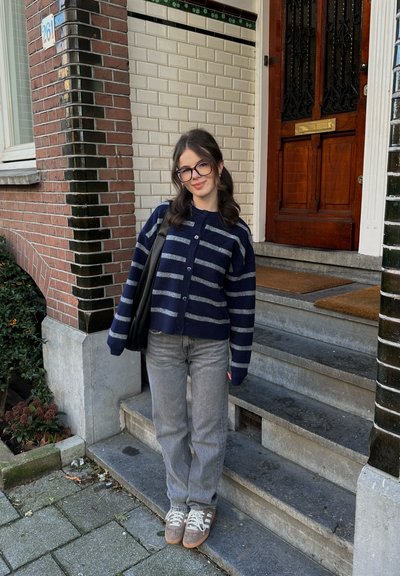 Cárdigan de punto a rayas en azul marino y gris sobre jeans grises. Zapatillas marrón claro con cordones blancos. De pie en unos escalones de concreto con fondo de pared de ladrillo.