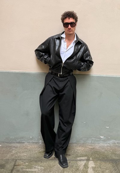 Hombre con chaqueta de cuero negra, camisa blanca, pantalones negros de pierna ancha, botas negras y grandes gafas de sol, apoyado contra una pared bicolor.