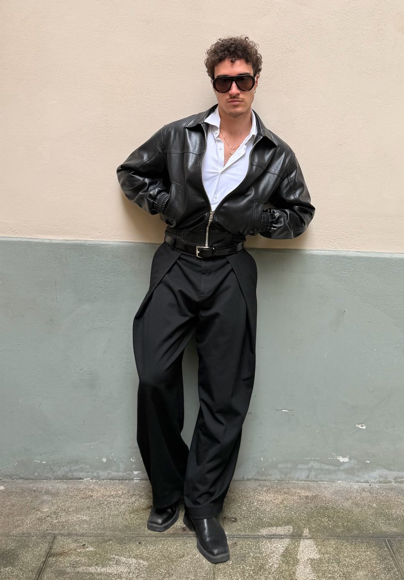 Άνδρας με μαύρο δερμάτινο μπουφάν, λευκό πουκάμισο, μαύρο παντελόνι φαρδιάς γραμμής, μαύρες μπότες και μεγάλα γυαλιά ηλίου, ακουμπά σε διχρωμικό τοίχο.