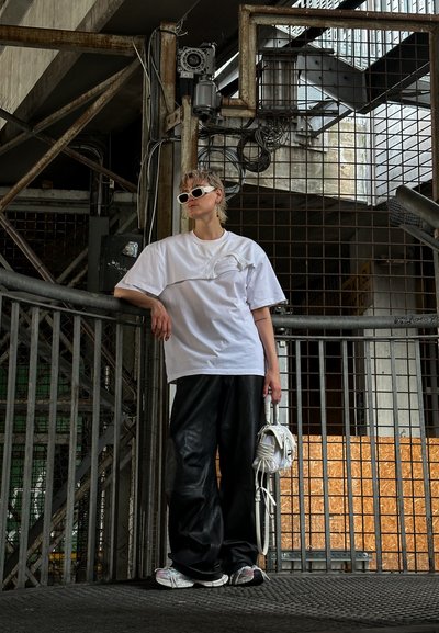 Camiseta blanca de gran tamaño con un diseño texturizado, pantalones de cuero negros sueltos y zapatillas multicolores, sosteniendo una bolsa blanca en un entorno urbano desgastado.