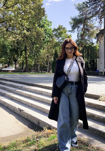 Abrigo negro largo, camiseta blanca, vaqueros anchos azules, bolso negro en forma de corazón y zapatillas negras. Árboles verdes y escaleras en el fondo.
