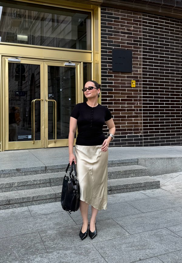 Femme avec des lunettes de soleil noires, un haut noir, une jupe en satin beige et des talons noirs, tenant un sac à main noir devant un bâtiment aux portes dorées.