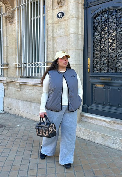 Mujer vestida con chaleco gris y pantalones deportivos con gorra beige, sosteniendo un bolso estampado de leopardo, de pie en la acera junto a un edificio de piedra y una puerta azul con el número 99.