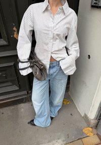 Fehér és világoskék csíkos hosszú ujjú ing, világoskék, bőszárú farmerrel párosítva. Fekete bőrtáska lóg a karon.