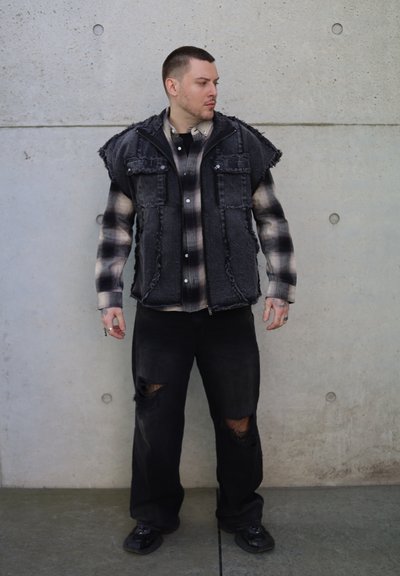 Chaleco de mezclilla negro con bordes deshilachados sobre una camisa de franela a cuadros en blanco y negro, combinado con pantalones negros desgastados y zapatos negros.