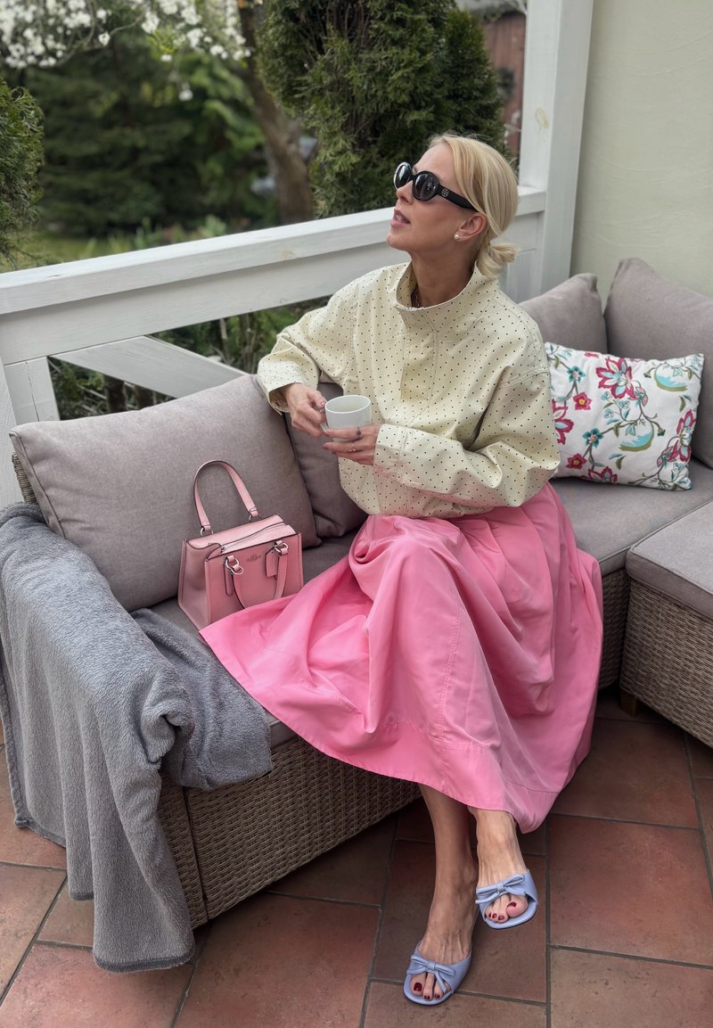 Femme portant des lunettes de soleil assise sur un canapé d'extérieur, vêtue d'une veste crème à pois, d'une jupe rose, de sandales bleues, tenant une tasse blanche.