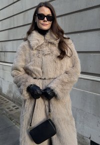 Manteau en fausse fourrure beige clair avec un col large, accompagné de gants en cuir noir et d'un petit sac bandoulière noir. Texture lisse.