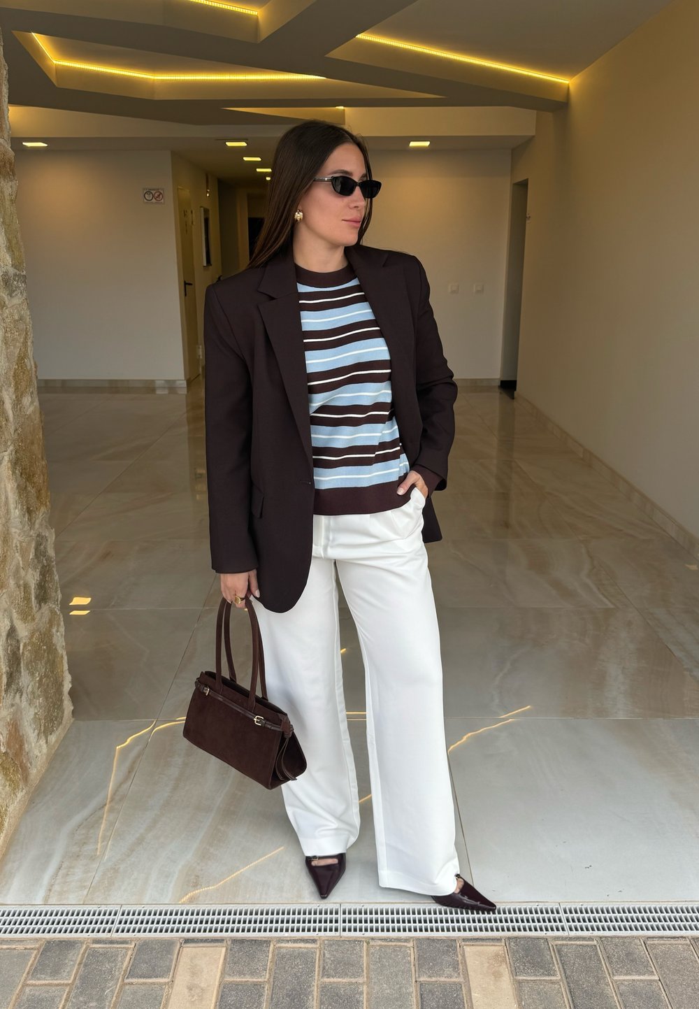 Blazer marrone scuro sopra un maglione a righe blu e marrone, pantaloni bianchi a gamba larga, tacchi marroni appuntiti e una piccola borsa marrone scuro.