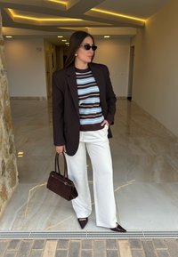 Blazer marrone scuro sopra un maglione a righe blu e marrone, pantaloni bianchi a gamba larga, tacchi marroni appuntiti e una piccola borsa marrone scuro.