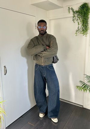 Hombre con gafas llevando una chaqueta bomber verde, vaqueros azules anchos, zapatillas blancas y llevando un bolso marrón al hombro en una habitación blanca con plantas.