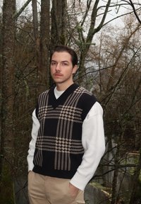 Pull sans manches à motif pied-de-poule noir et marron porté sur une chemise blanche à manches longues, associé à un pantalon beige dans un cadre forestier.