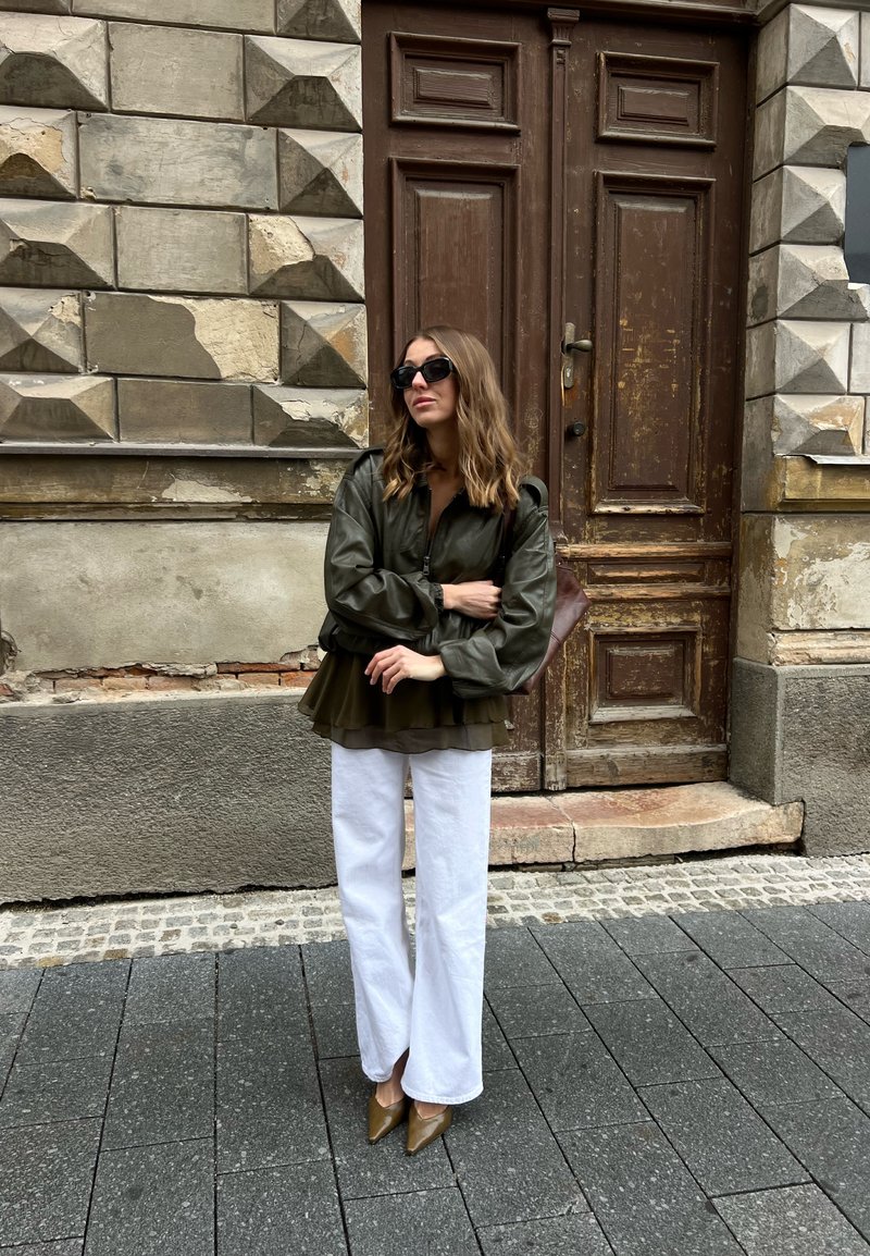 Frau mit Sonnenbrille, grüner Jacke, weißer Hose und braunen Schuhen, steht mit verschränkten Armen neben einer verwitterten Holztür und einer strukturierten Steinwand.