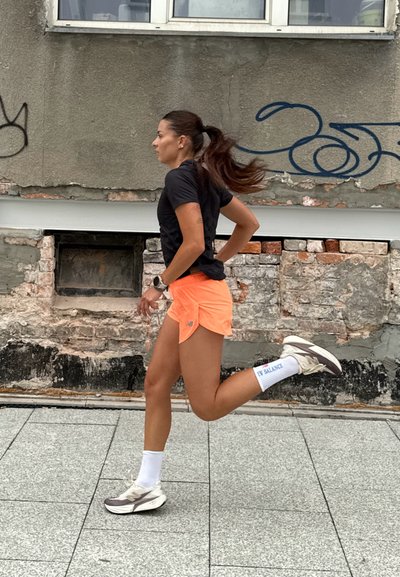 Atleta con una camiseta de manga corta negra, pantalones cortos neon naranjas y calcetines blancos con detalles azules, corriendo sobre un pavimento texturizado cerca de una pared de ladrillos.