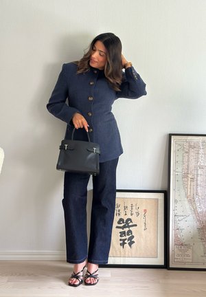 Chaqueta de mezcla de lana en azul marino con botones dorados, combinada con pantalones de mezclilla oscuros y tacones negros con tiras, sosteniendo un bolso de cuero negro.