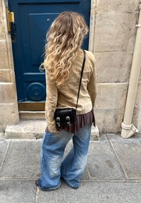 Vrouw met lang golvend blond haar, gekleed in een beige jack, brede blauwe jeans en een zwarte schoudertas, staande op stenen bestrating voor een blauwe deur.