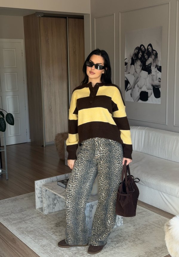 Maglione a righe oversize giallo e marrone; pantaloni a zampa di leopardo; borsa scura; scarpe basse; ambiente interno con decorazione minimalista.
