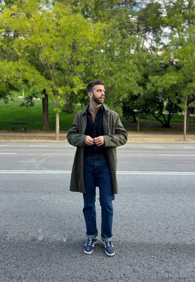 Hombre con un largo abrigo verde olivo sobre una camisa negra, jeans azules y zapatillas navy, de pie en una calle bordeada de árboles.