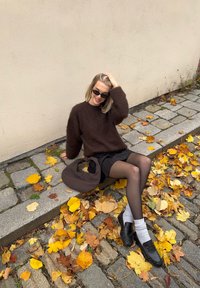 Suéter castanho fofinho, saia preta e mocassins pretos com meias cinzentas. Cercada por folhas amarelas de outono e um caminho de pedras texturizado.