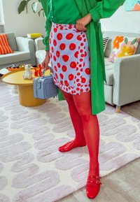 Grønn skjorte, mønstret skjørt med røde sirkler på lyseblått, røde strømpebukser, røde sko med spenner, holder en liten blå håndveske.