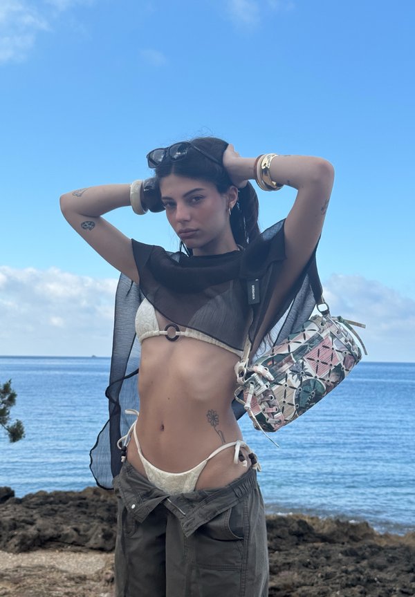 Młoda kobieta w białym bikini i przezroczystym czarnym topie pozująca z rękami za głową w pobliżu skalistego brzegu i spokojnego morza pod błękitnym niebem.