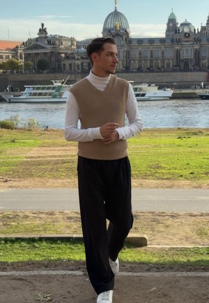 Chaleco de punto beige sobre un jersey de cuello alto blanco, combinado con pantalones plisados negros y zapatillas blancas. El fondo presenta un río y edificios históricos.