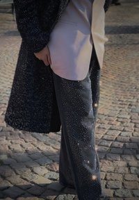 Palton texturat negru peste o cămașă cu butoni de culoare roz deschis. Pantalonii sunt închisi la culoare, având un model strălucitor de stele și dungi laterale contrastante.