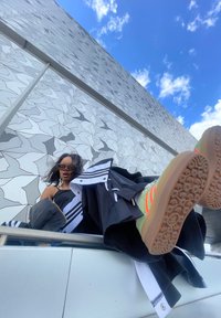 Veste noire et blanche zippée avec des rayures, superposée à une tenue noire fluide. Baskets colorées et à motifs avec une semelle texturée. Mur géométrique en arrière-plan.