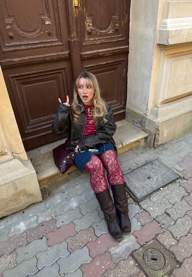 Jeune femme aux cheveux blonds portant une veste en cuir, une chemise rayée, une jupe en jean, des collants rouges en dentelle et des bottes, assise sur une marche en pierre près d'une porte en bois.