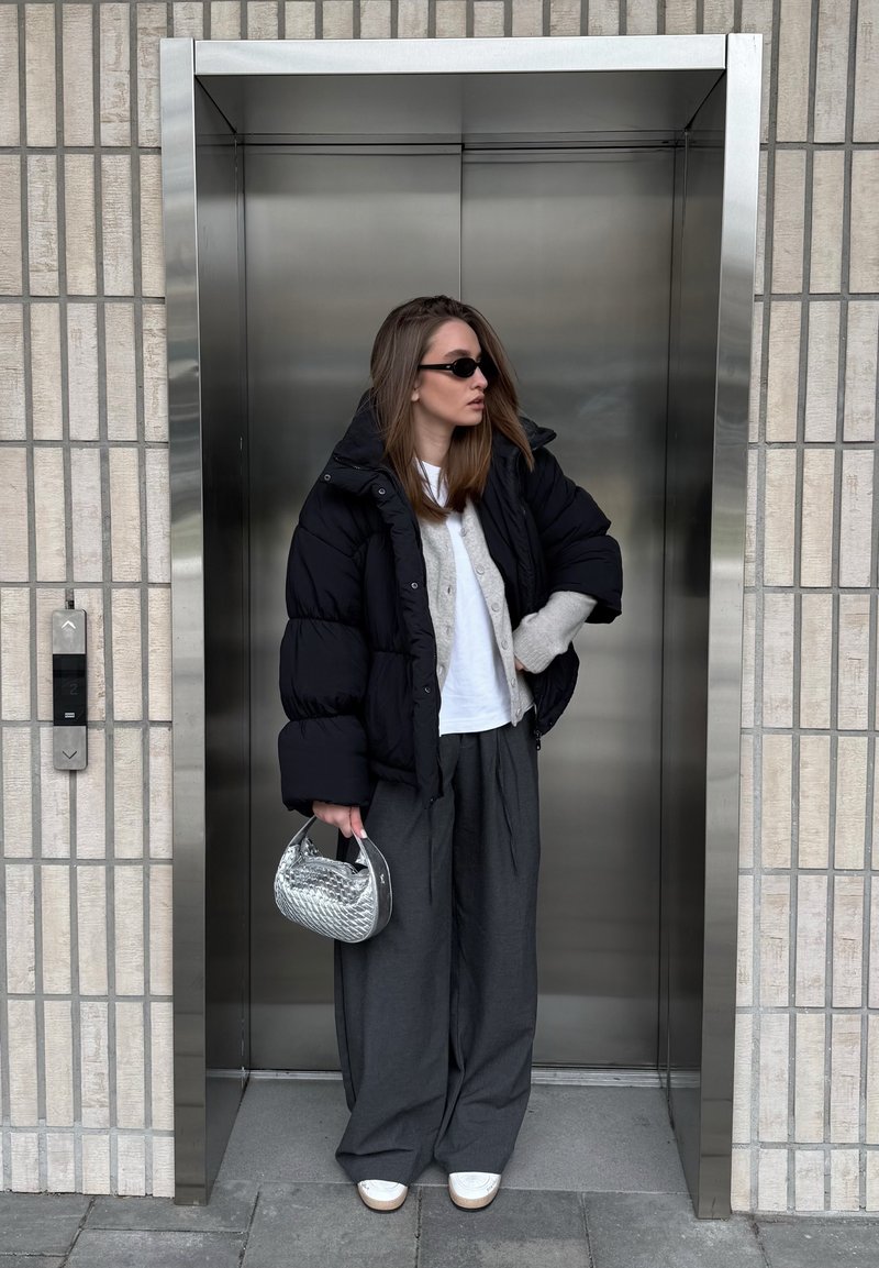 Frau trägt schwarze Steppjacke, graue Hose, weiße Turnschuhe, hält eine silberne Handtasche und steht vor geschlossenen Aufzügen aus Edelstahl.