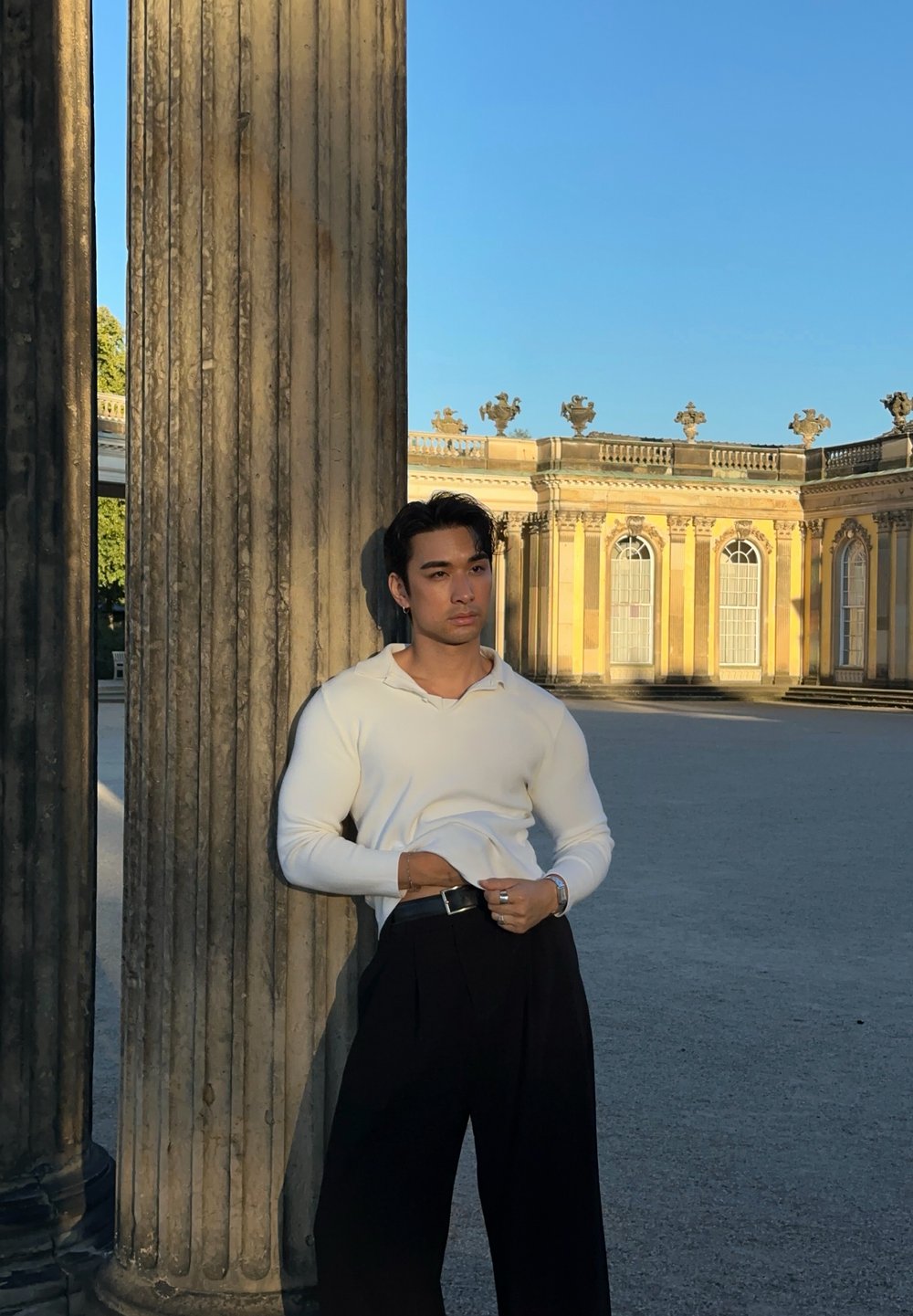 Junger Mann in weißem Hemd und schwarzer Hose, der sich an einer großen Steinsäule lehnt, mit einem historischen gelben Gebäude und klarem blauen Himmel im Hintergrund.