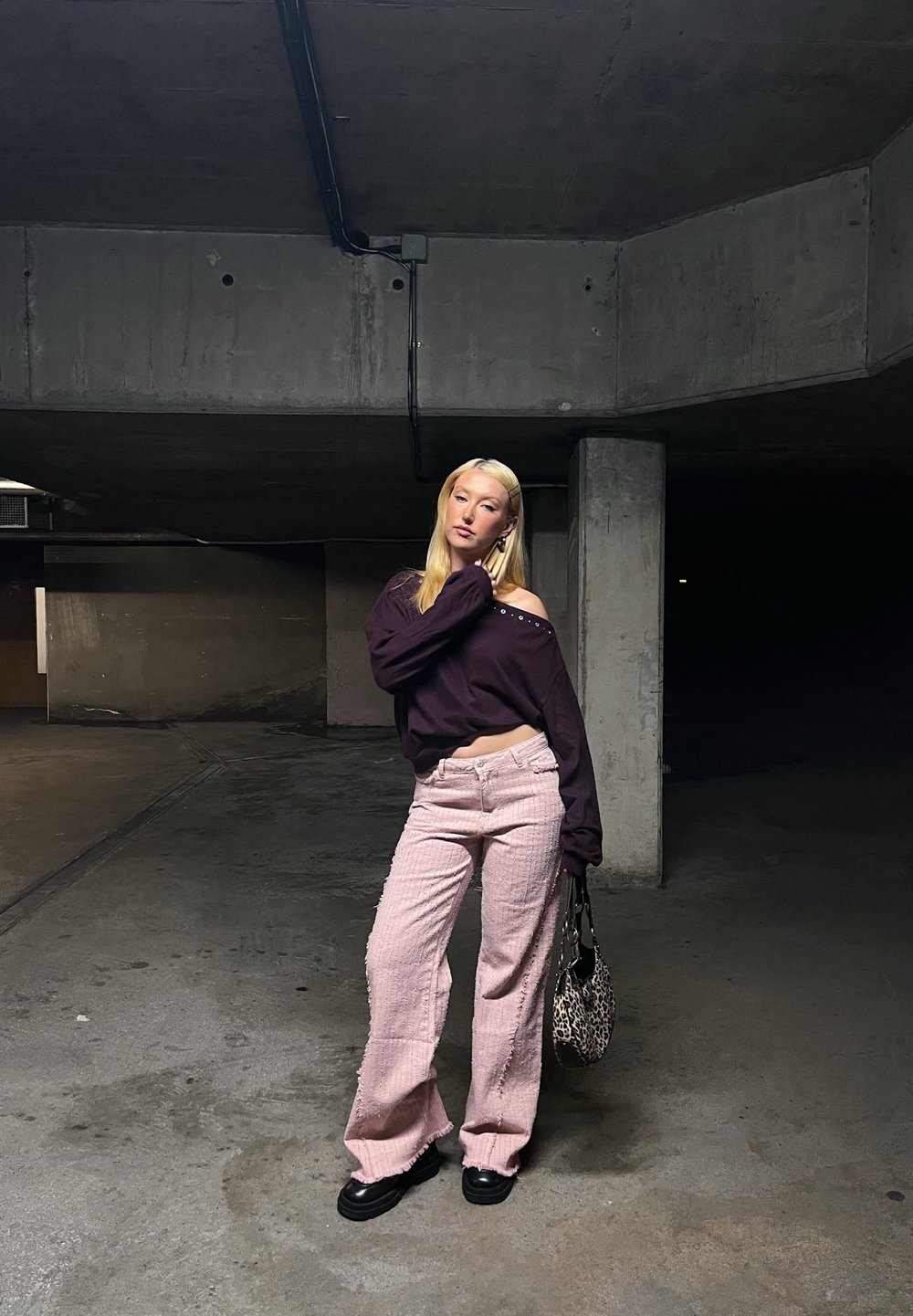 Överdimensionerad lila skjorta med off-the-shoulder-detalj, matchat med ljusrosa texturerade byxor och svarta plattformsskor; håller en leopardmönstrad väska.