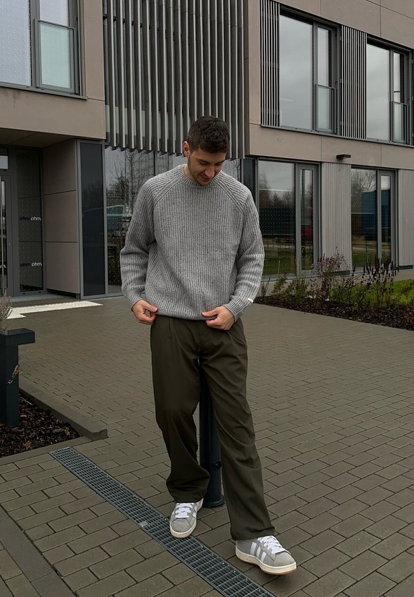Grauer, gerippter Pullover, olivgrüne Hose und hellgraue Turnschuhe. Steht auf gefliestem Boden in der Nähe eines modernen Gebäudes mit großen Fenstern.
