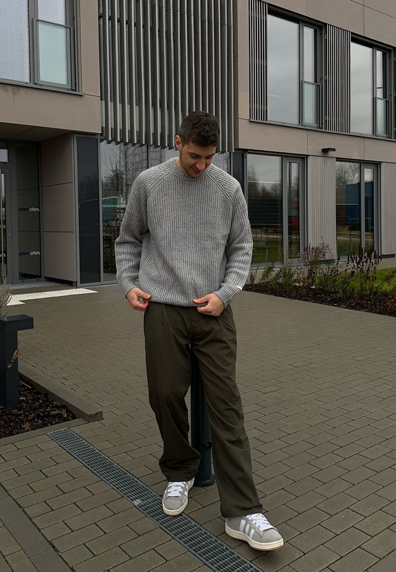 Pull gris côtelé, pantalon vert olive et baskets gris clair. Debout sur un sol carrelé près d'un bâtiment moderne avec de grandes fenêtres.