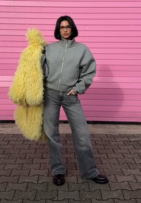 Grå zip-up sweatshirt, grå jeans med vida ben och svarta platåskor. Håller ett fluffigt gult täckjacka. Rosa bakgrund med horisontella linjer.