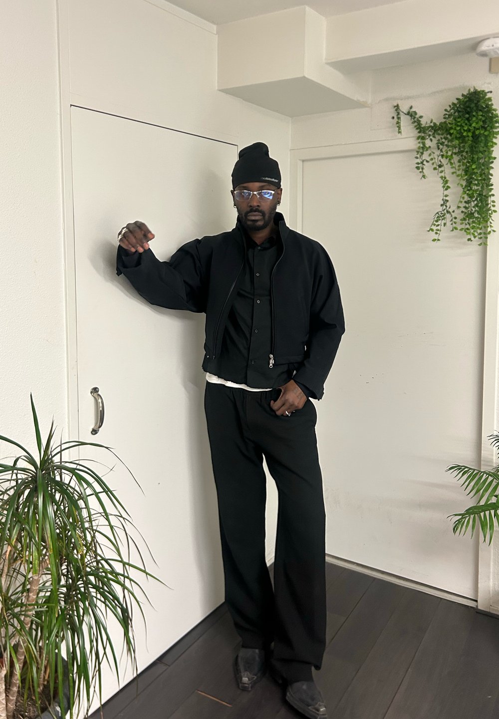 Veste noire courte portée sur une chemise noire à boutons, accompagnée d'un pantalon noir ample. Lunettes transparentes et bonnet noir. Plantes en arrière-plan minimalistes.