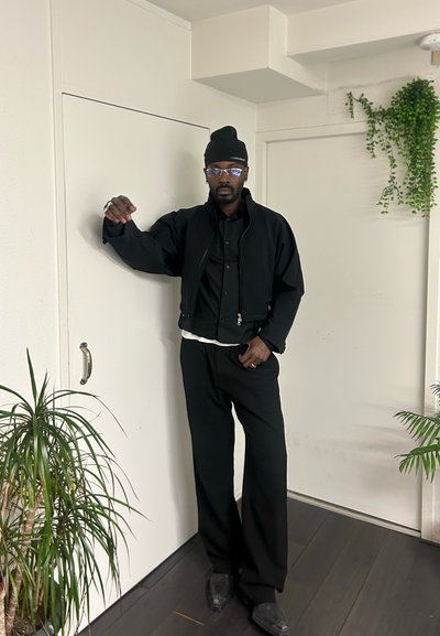 Chaqueta corta negra sobre una camisa negra abotonada, combinada con pantalones negros anchos. Gafas transparentes y un gorro negro. Fondo minimalista con plantas.