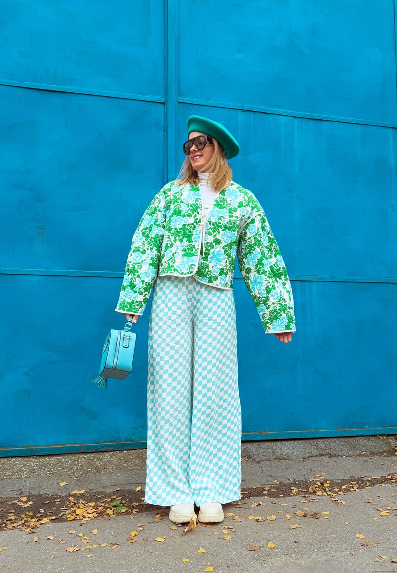 Veste matelassée florale verte, col roulé blanc, pantalon large à carreaux bleus et blancs, béret vert et sac à main bleu ciel.