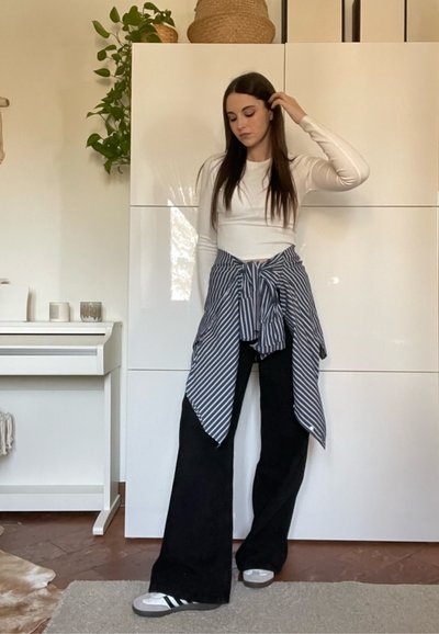 Mujer con cabello largo y oscuro, vestida con una camiseta blanca de manga larga, pantalones anchos negros y una camisa de rayas atada a la cintura, de pie en una habitación con plantas y gabinetes blancos.
