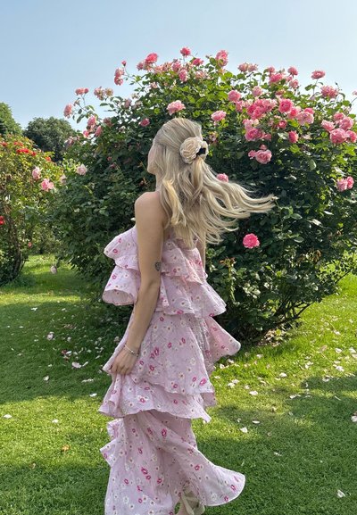 Vestido estampado en rosa con capas con volantes y tejido texturizado, complementado con un accesorio para el cabello floral, en un entorno de jardín.