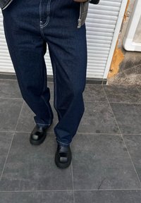 Temno modri kavbojki z belim šivom, oblečeni z črnimi natikači s debelim podplatom in gladko površino.